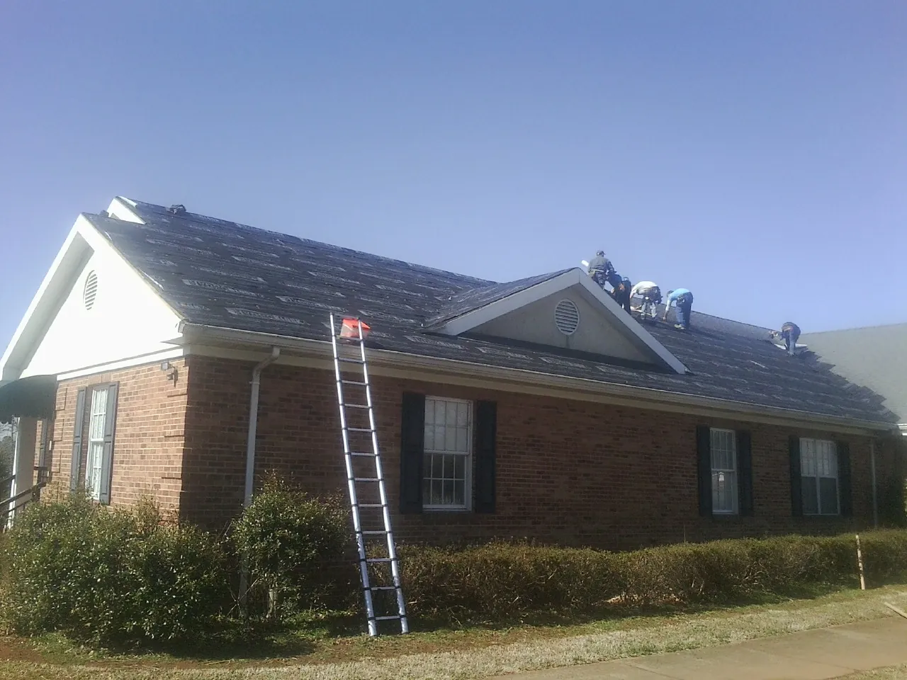 Crew tearing shingles off a brick ranch roof with a ladder set — Metal Roof Repair work by our crew in Wanatah