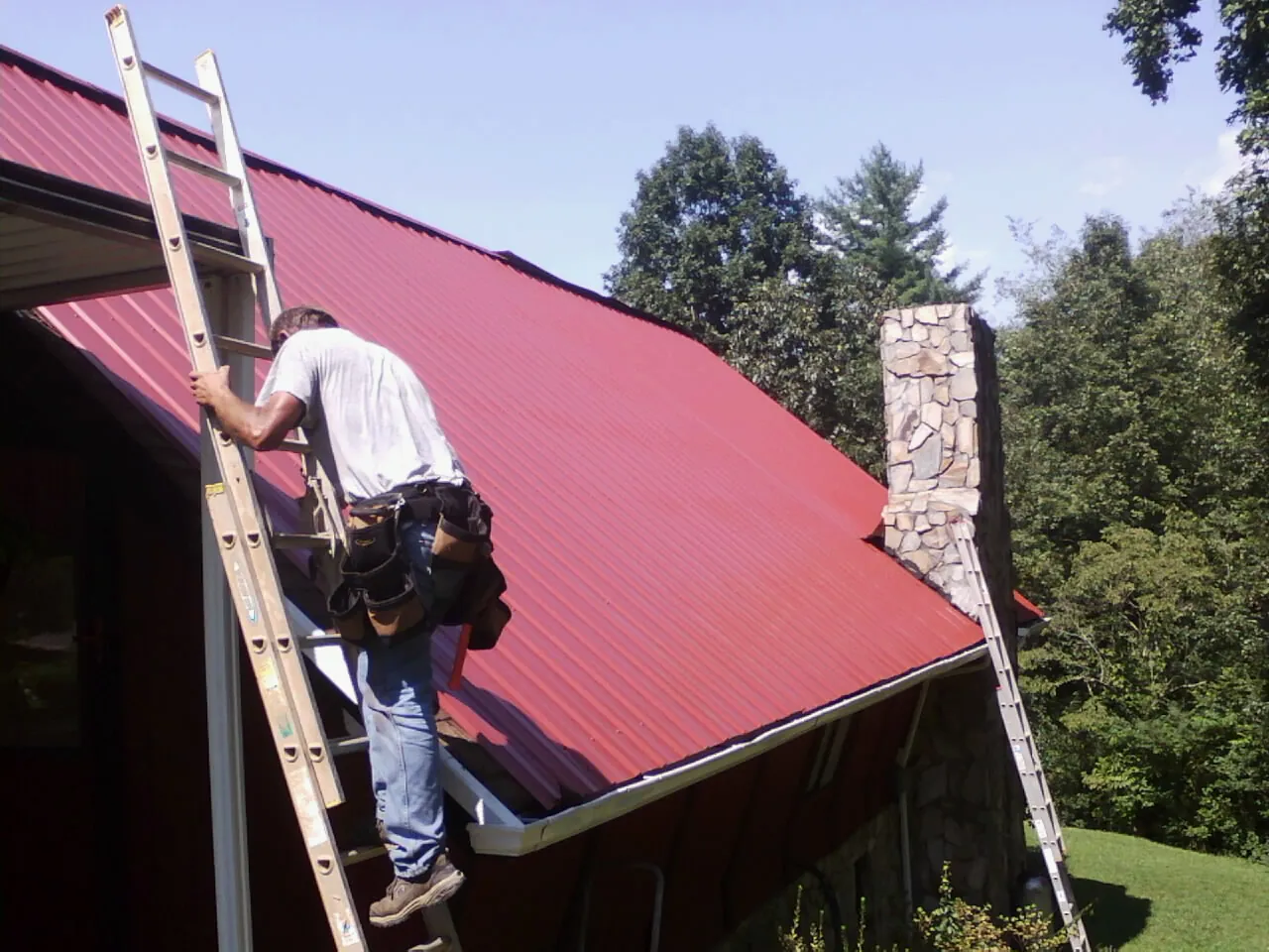 Roofer on a ladder beside a red metal roof and stone chimney — Soffit & Fascia Repair by Roofing Brothers in Wanatah